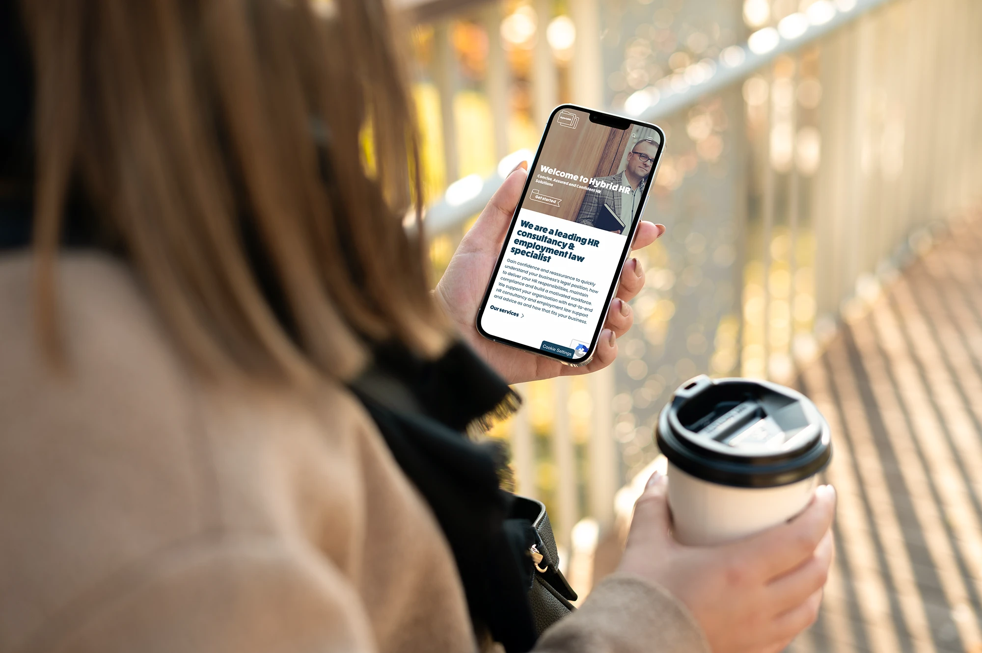 Person holding smartphone and coffee cup outdoors with blurred background.