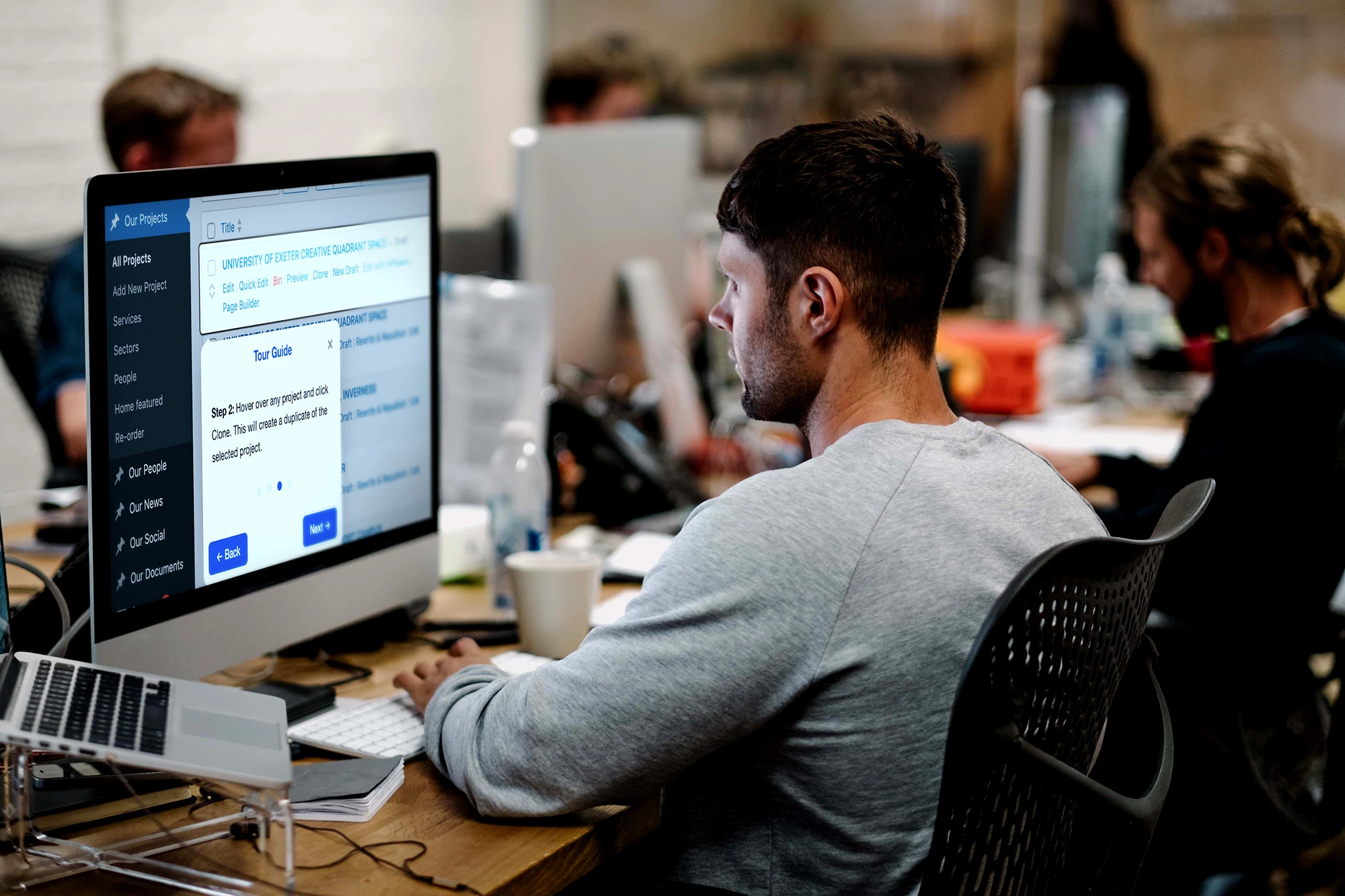 Young man working on a computer in a busy office environment with colleagues.