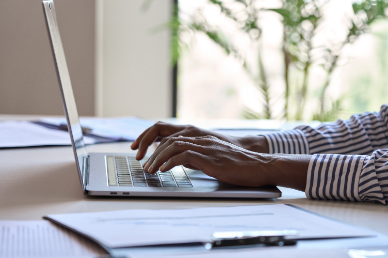 Person typing on a laptop with documents on a desk in a bright office.