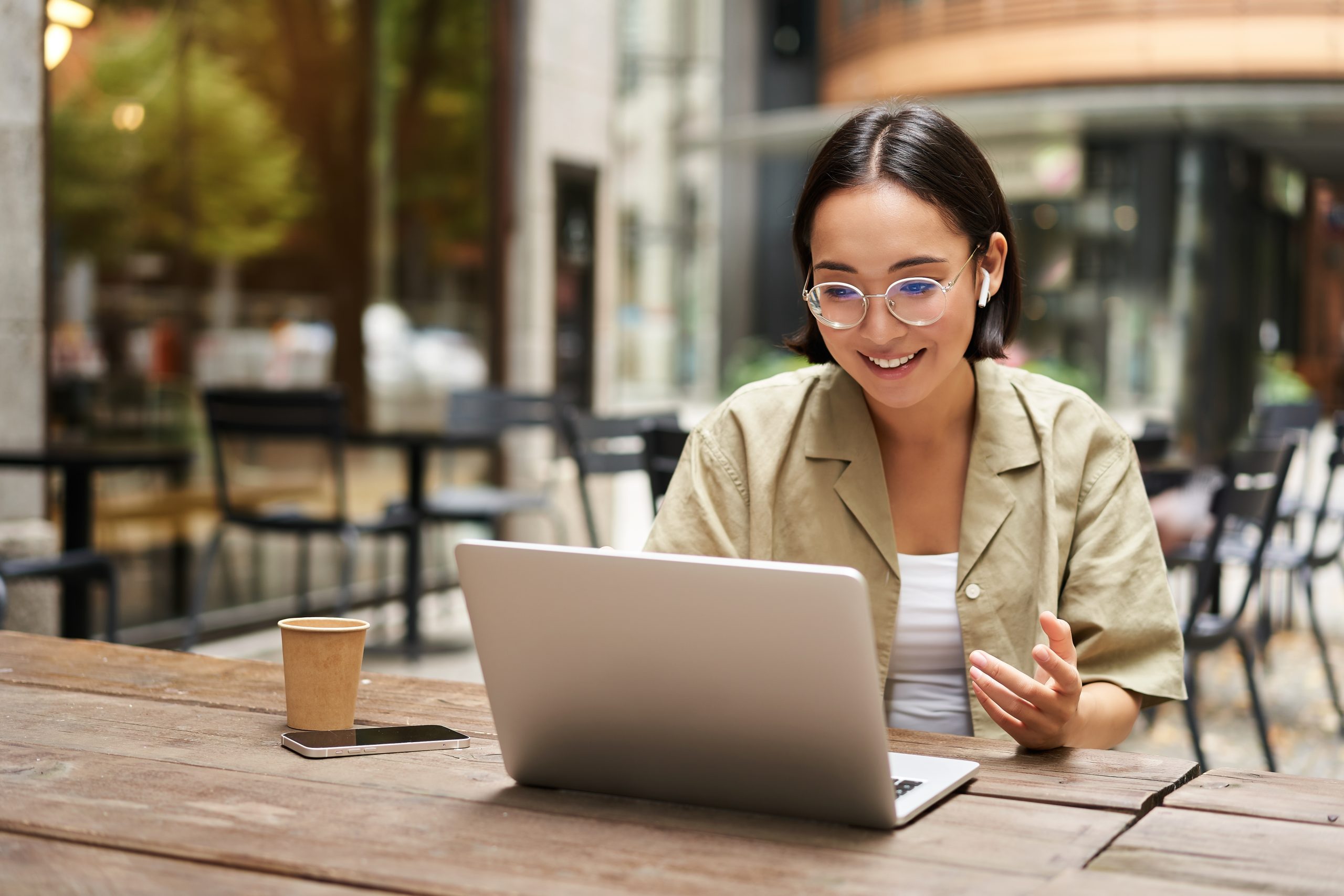 Woman working on a laptop outdoors at a cafe with a coffee cup nearby.